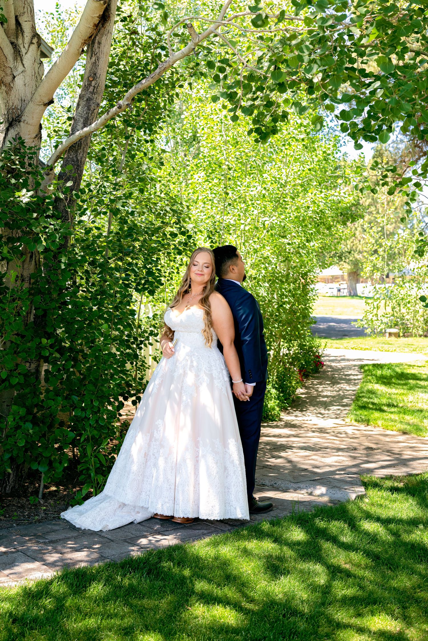 Bride in white gown and groom in navy suit hold hands on a tree-lined garden path on their wedding day.