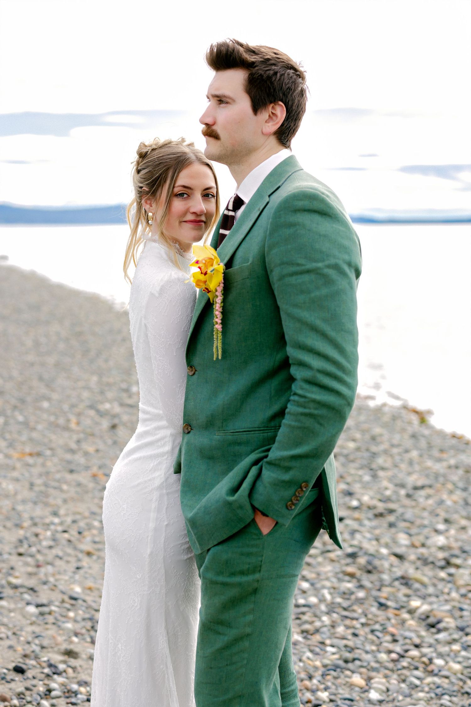 Bride and groom pose on rocky beach, groom wearing green suit with yellow boutonniere, bride in white dress.