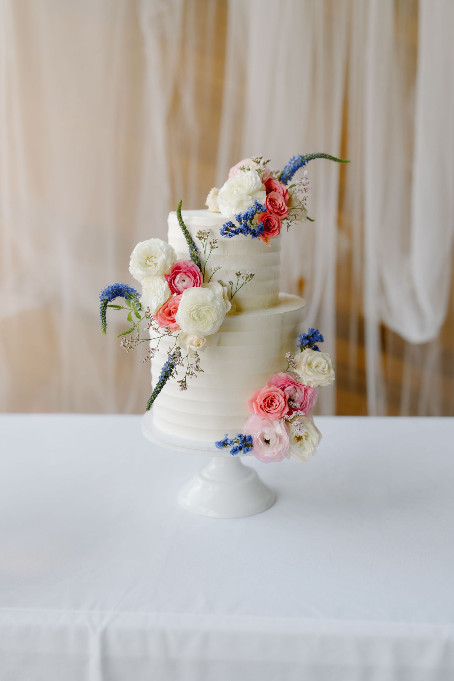 Elegant two-tier white wedding cake with pink, white, and blue floral decorations on a pedestal stand.