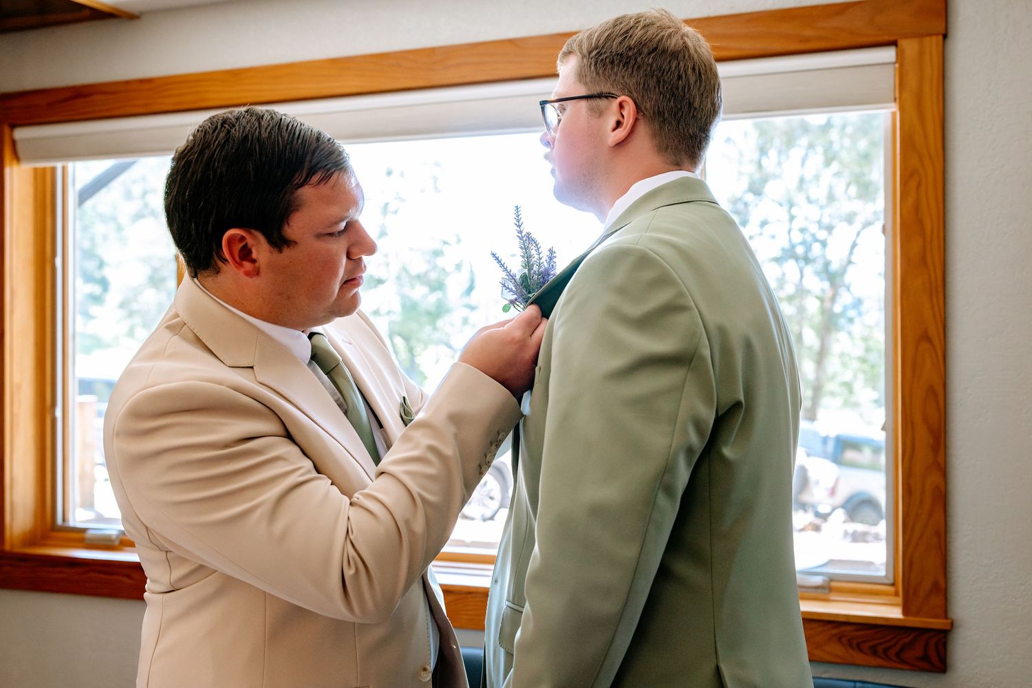 Two men in sage green suits, one pinning a boutonniere on the other before a wedding ceremony.