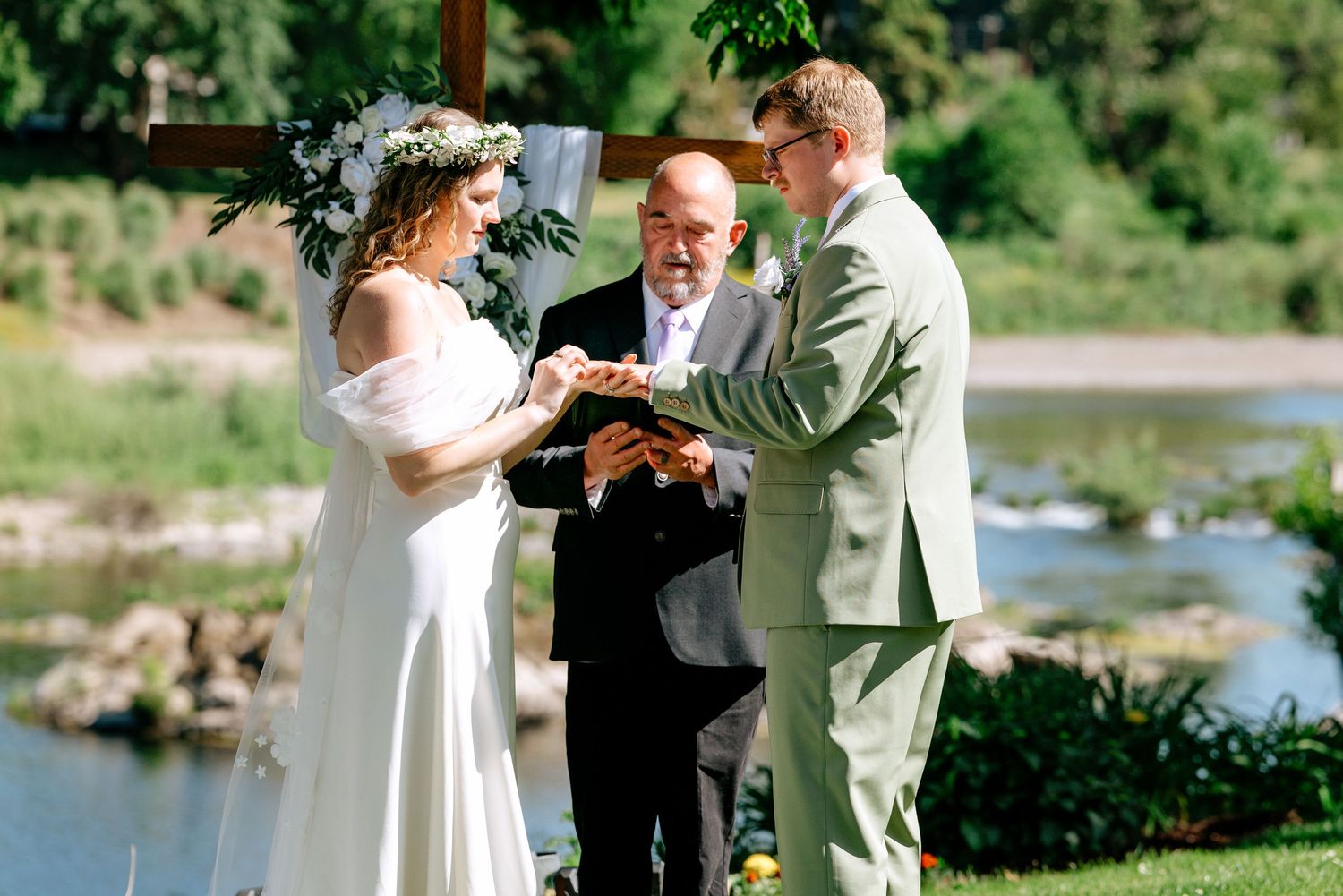 Bride and groom exchange rings outdoors with officiant, river backdrop, bride wearing floral crown, groom in sage suit.
