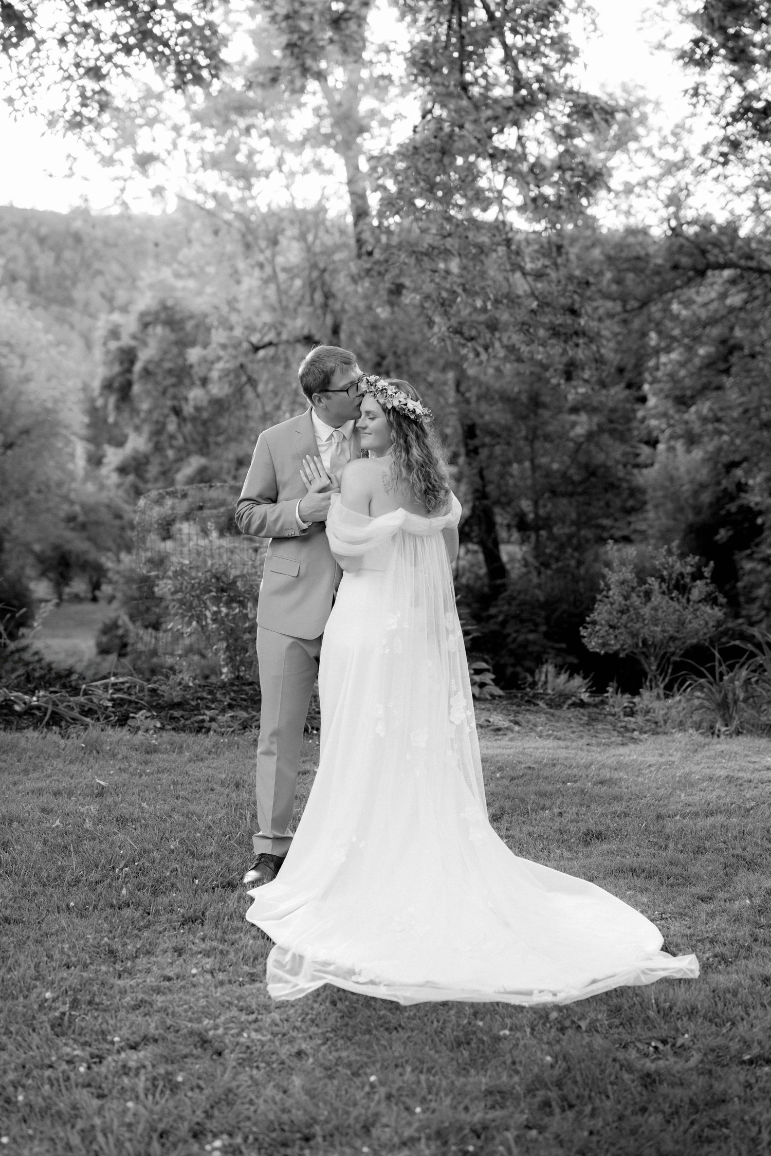 Black and white portrait of bride in flowing gown with floral crown embracing groom in suit outdoors.