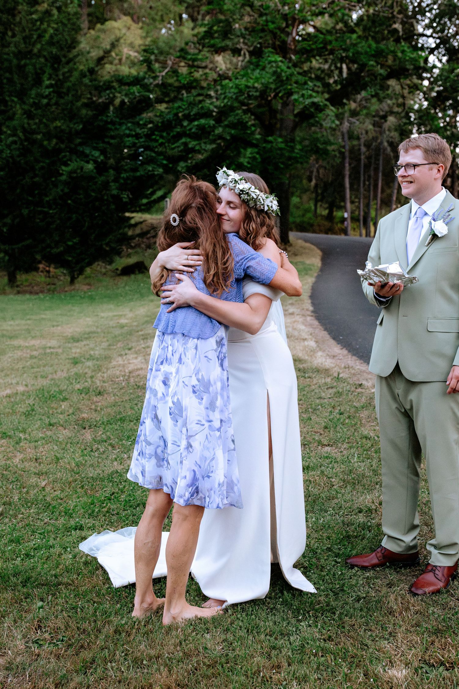 Bride in white gown and floral crown hugs guest in blue floral dress as groom in sage suit watches outdoors.