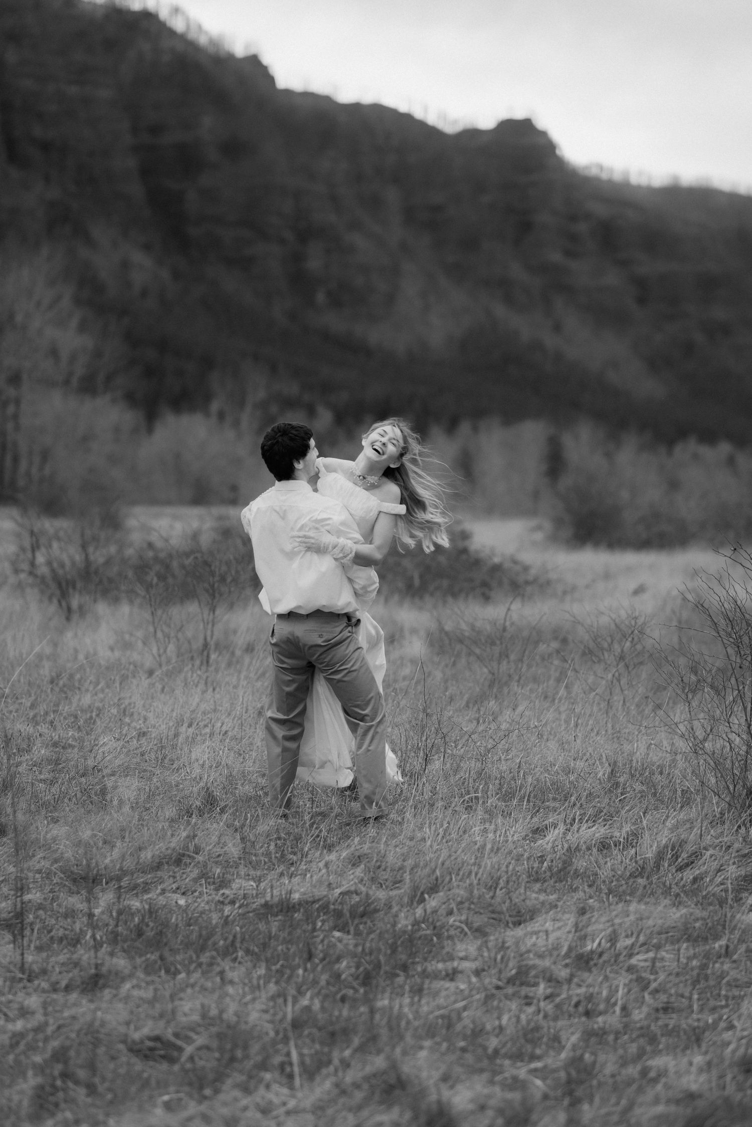 Black and white photo of a couple dancing playfully in a field with hills behind them.