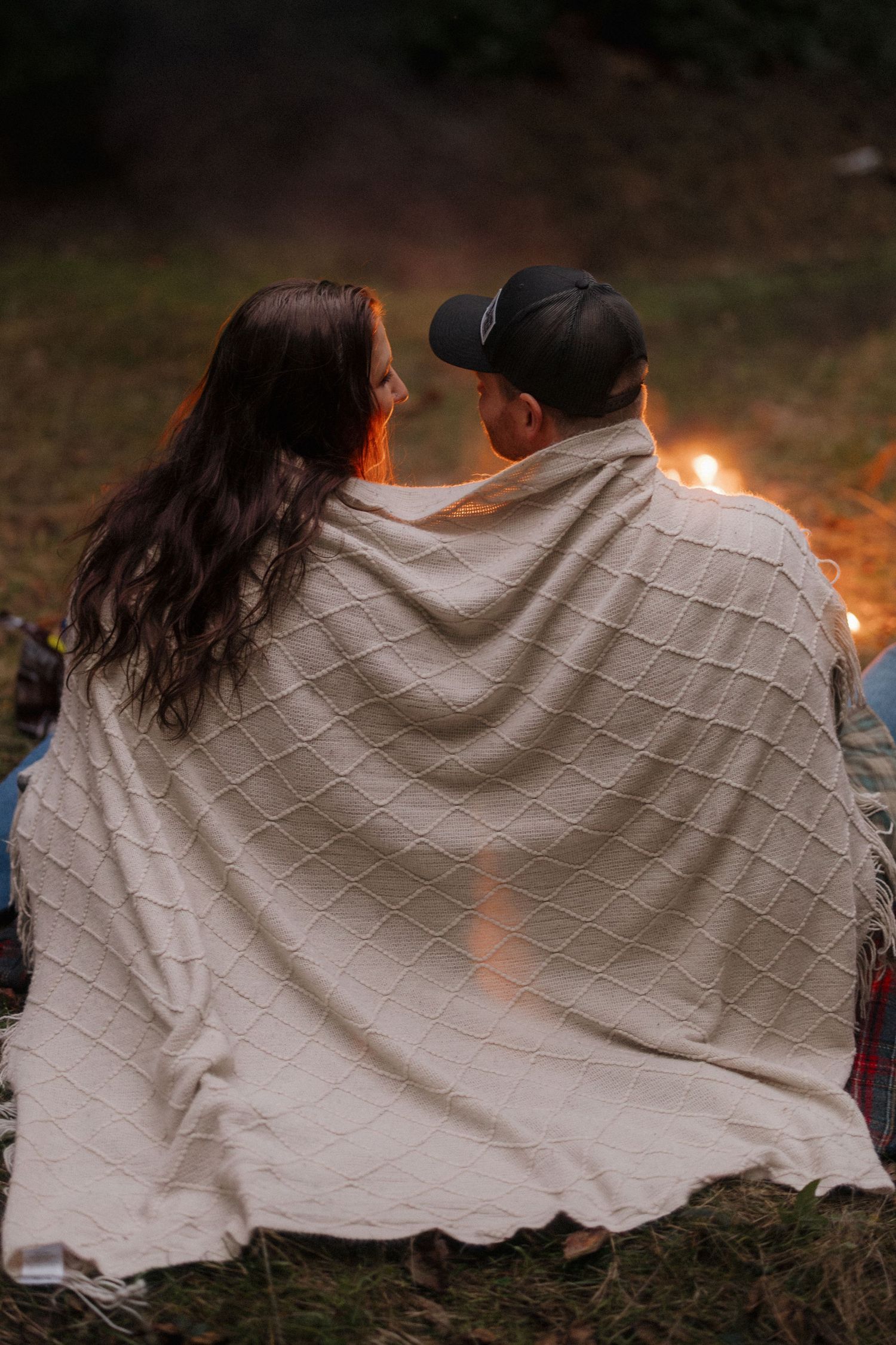 Couple wrapped in a cozy white blanket sitting by a campfire under the evening sky.