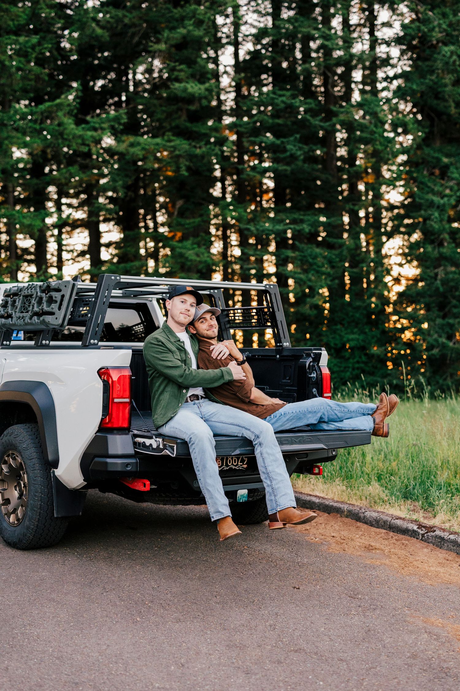 A couple sits in the back of a pickup truck parked on a forest road at sunset with evergreen trees in the background.