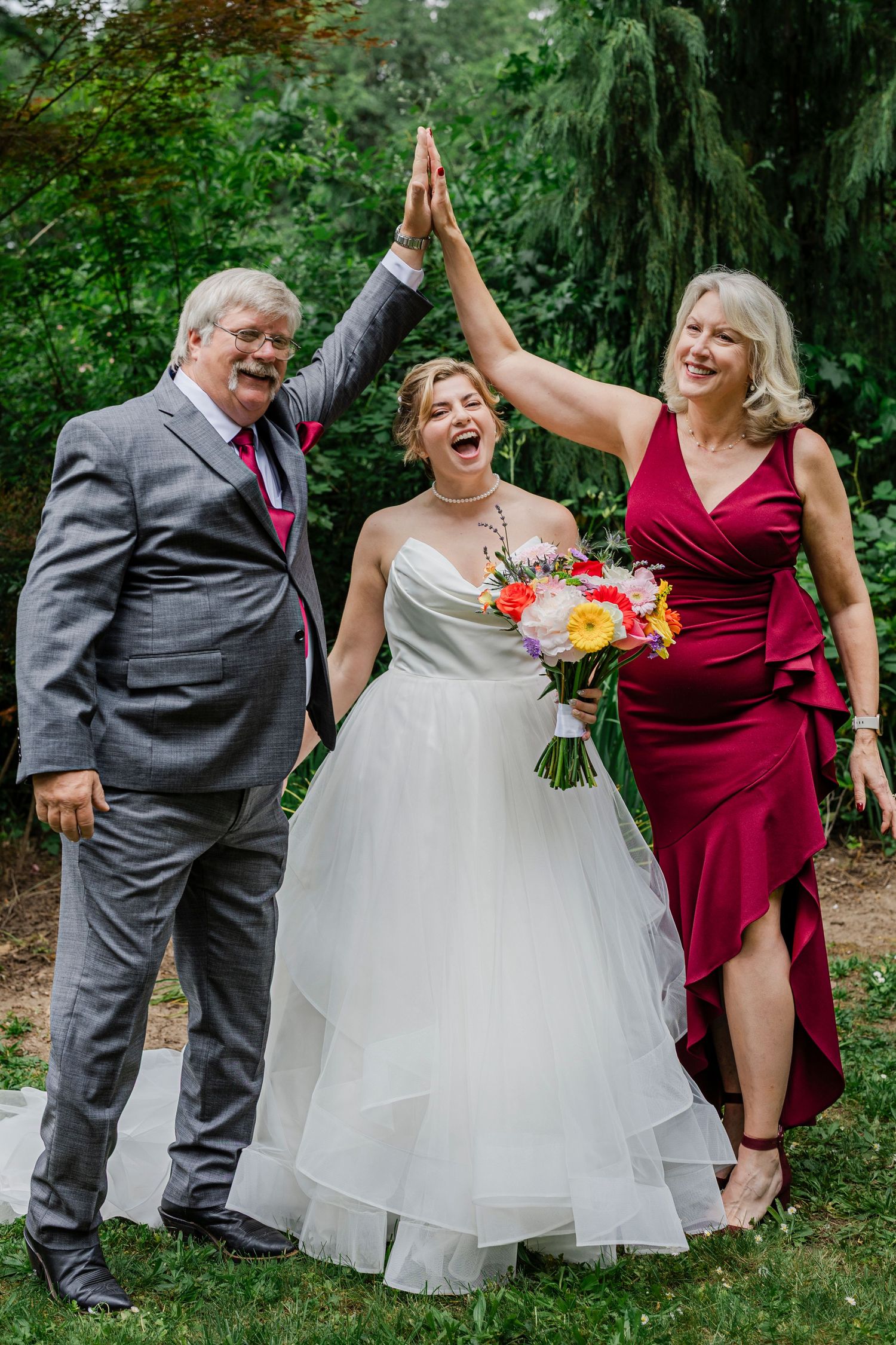 A wedding party celebrates with a high-five in a lush garden setting with colorful bouquet.