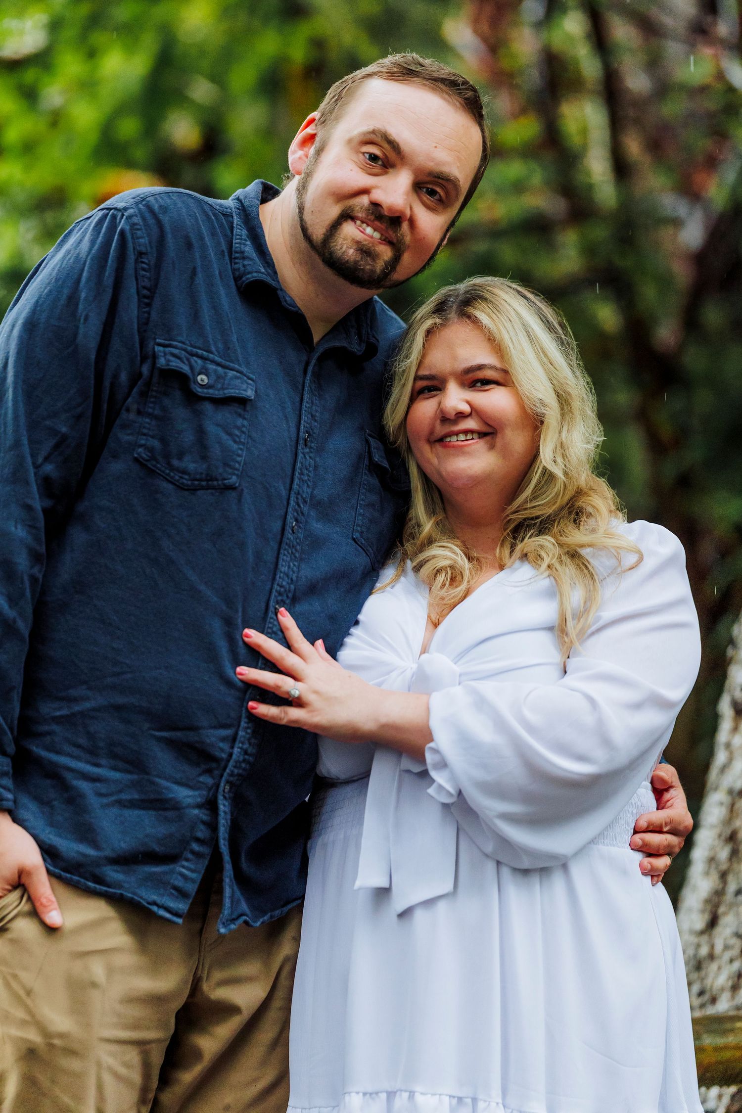 A couple poses together outdoors wearing a navy blue shirt and white dress against a green natural background.