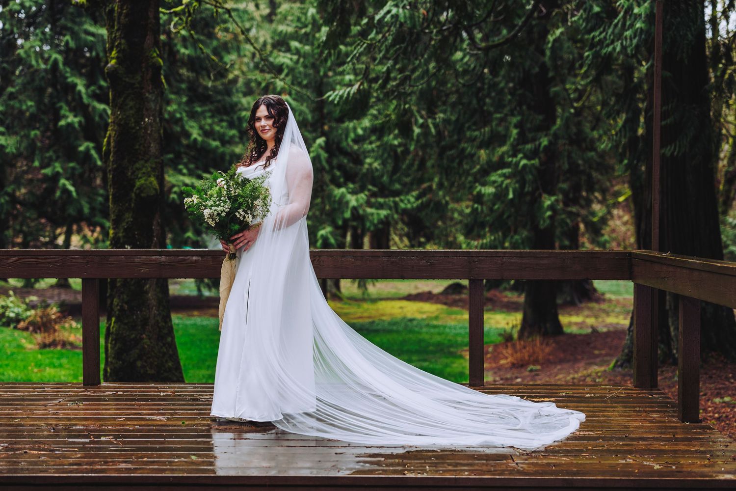 A white wedding gown with a flowing cathedral veil stands elegantly on a wooden bridge in a lush forest setting.