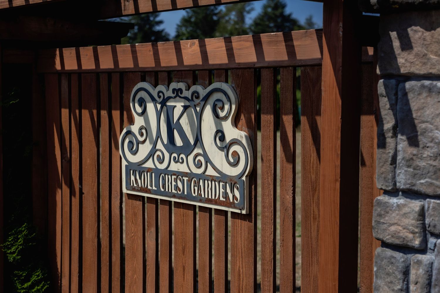 Rustic wooden fence with decorative iron scrollwork and stone pillars frames an entrance to a garden path.