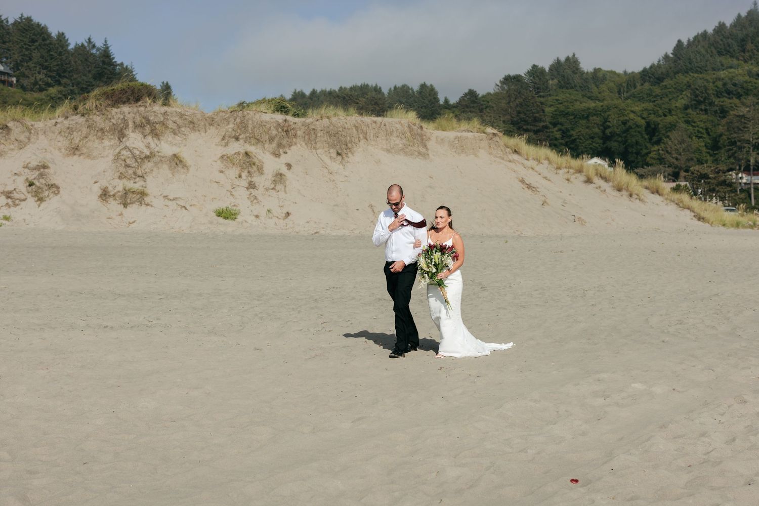 A couple poses for a wedding photo on a sandy beach with dunes and forest in the background.