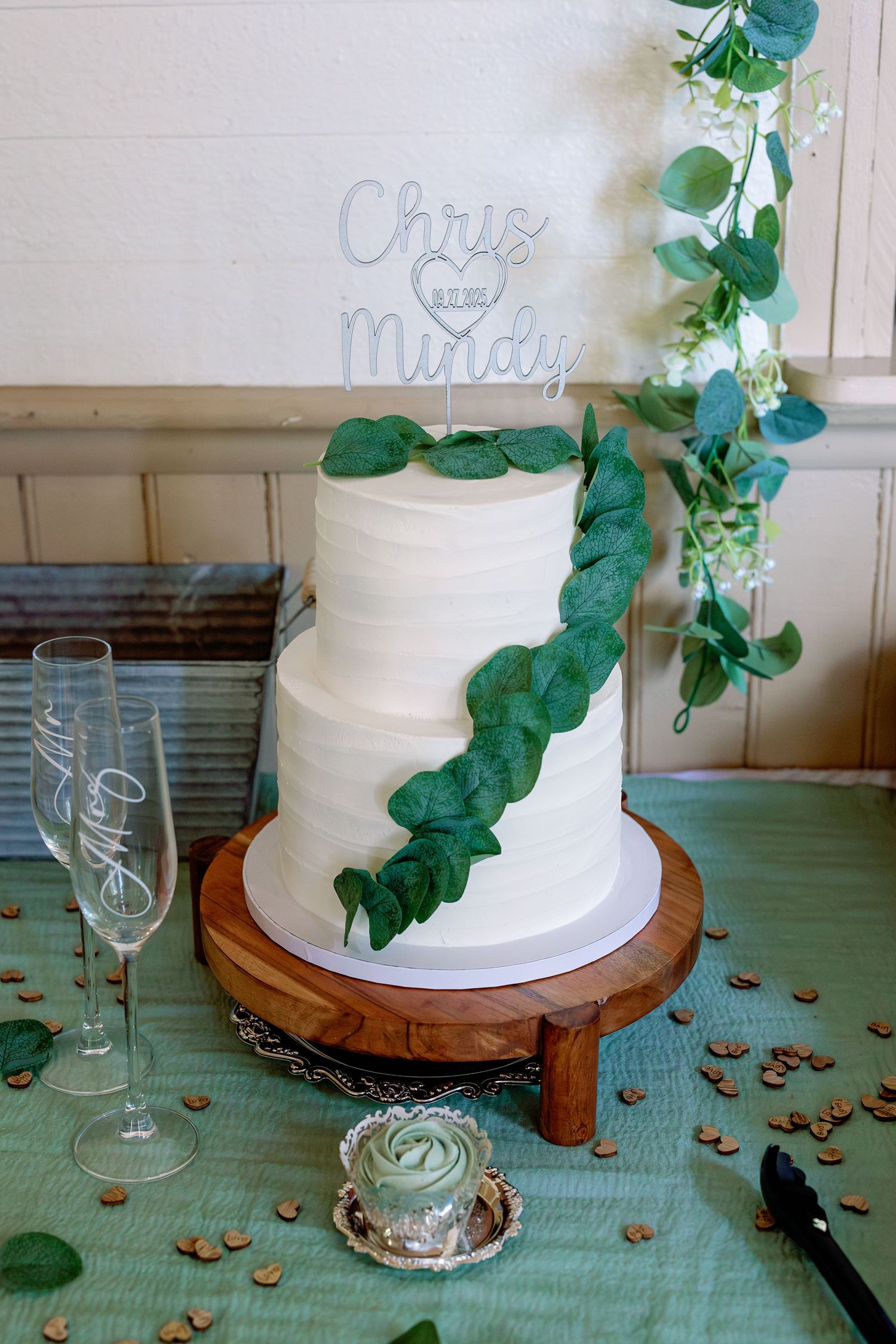 White wedding cake with eucalyptus garland displayed on wooden stand with wine glasses and turquoise tablecloth.