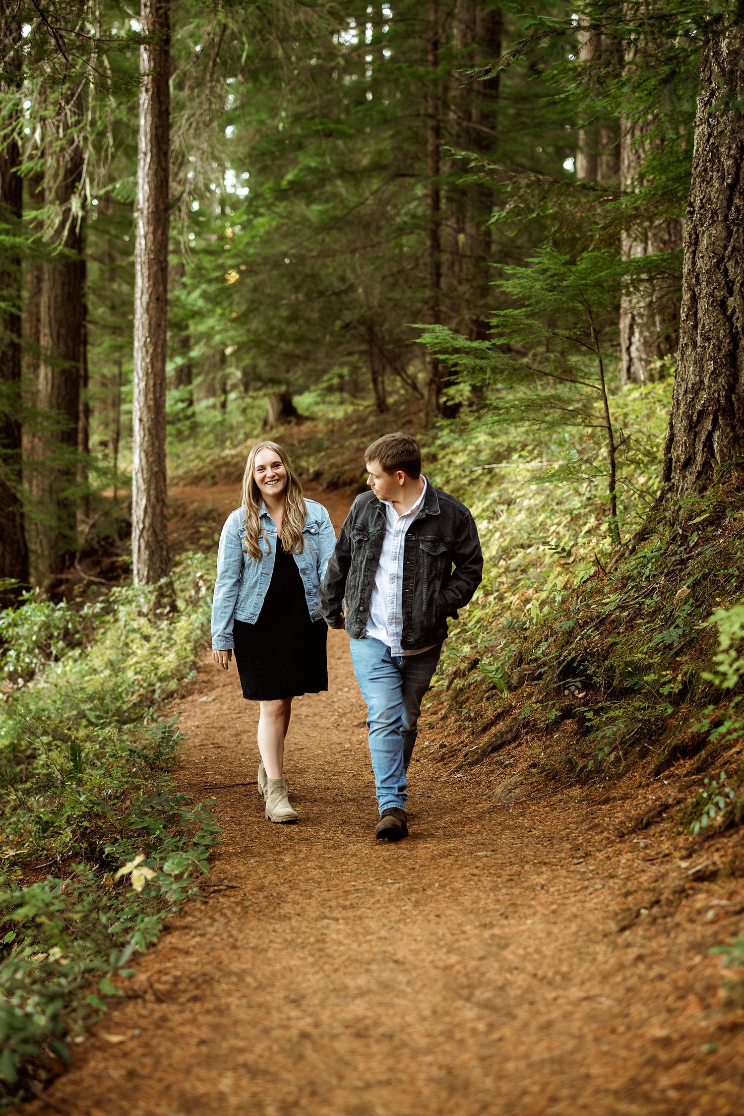 Two people walking hand in hand on a winding dirt trail through a dense forest.