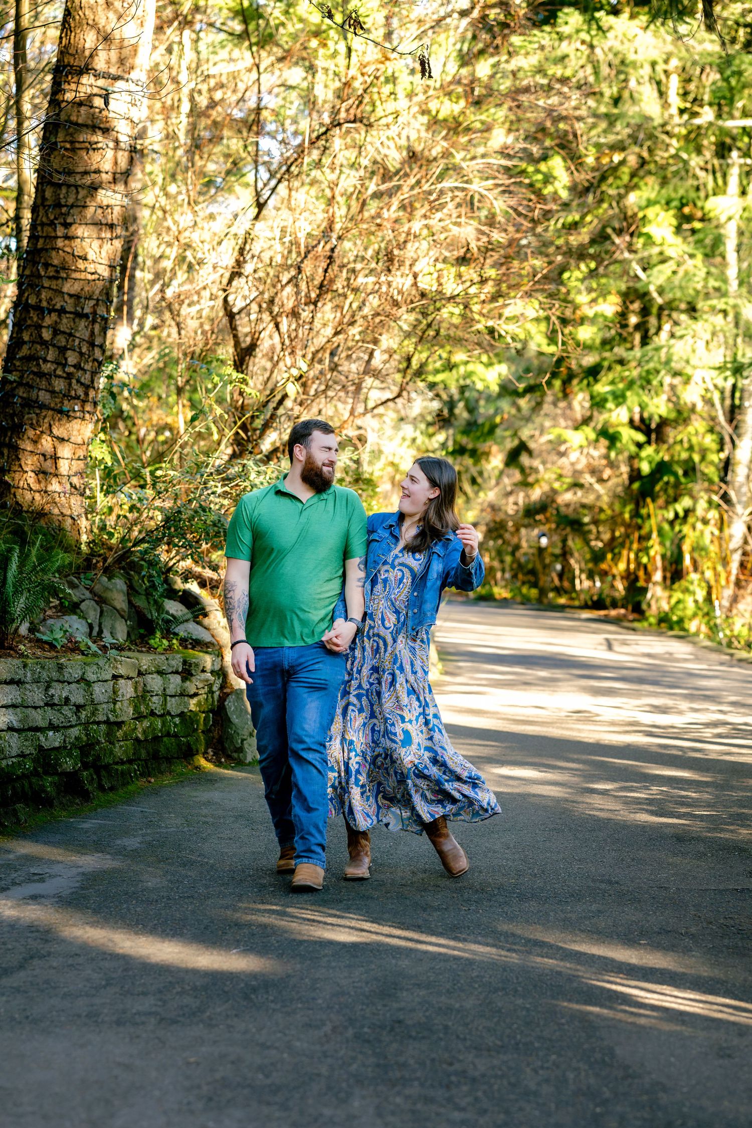 A couple sits together on a stone wall along a sunlit path surrounded by lush greenery and trees.