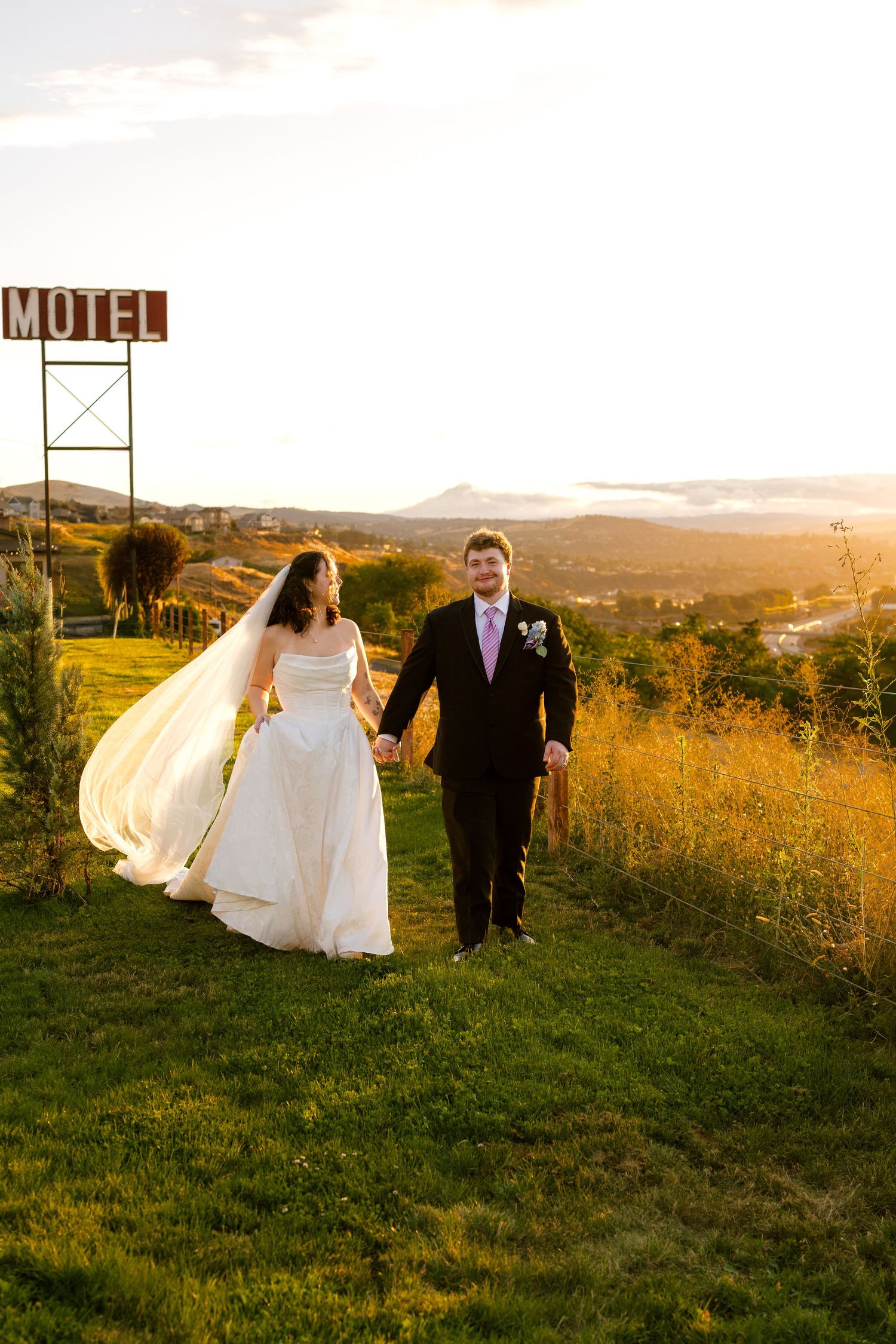 Newlyweds walking through golden field at sunset with dress flowing in wind.