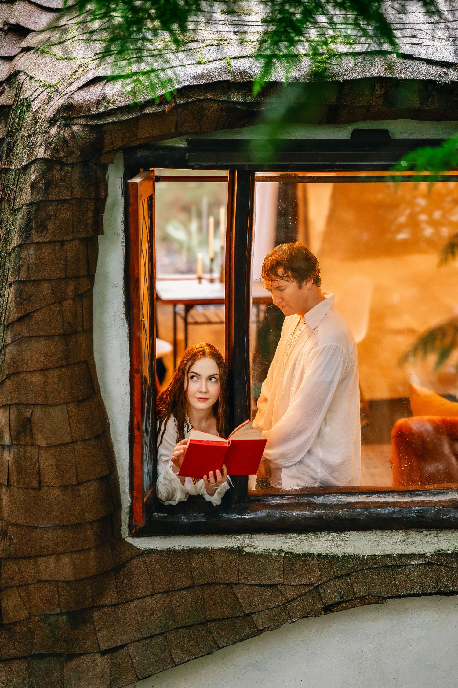 Through a window frame, two people sit together in a warmly lit interior with tree branches visible outside.