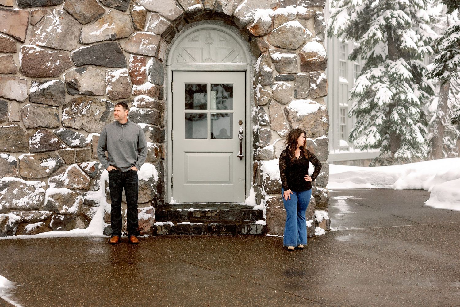 Two people stand casually by a grey door framed by a rustic stone archway with snow.