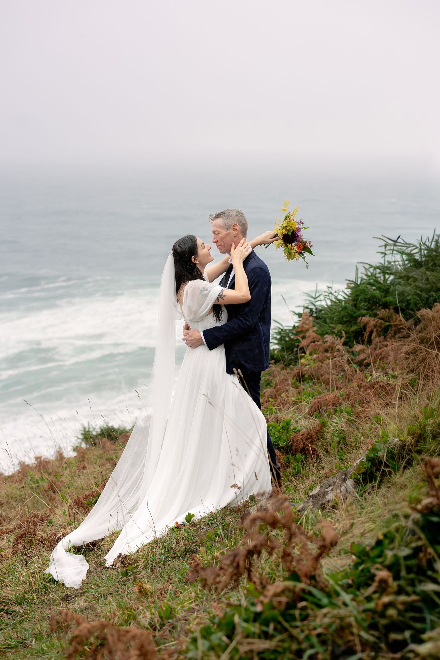 A romantic moment captured on a dramatic coastal cliff overlooking the ocean with flowing white dress in the wind.