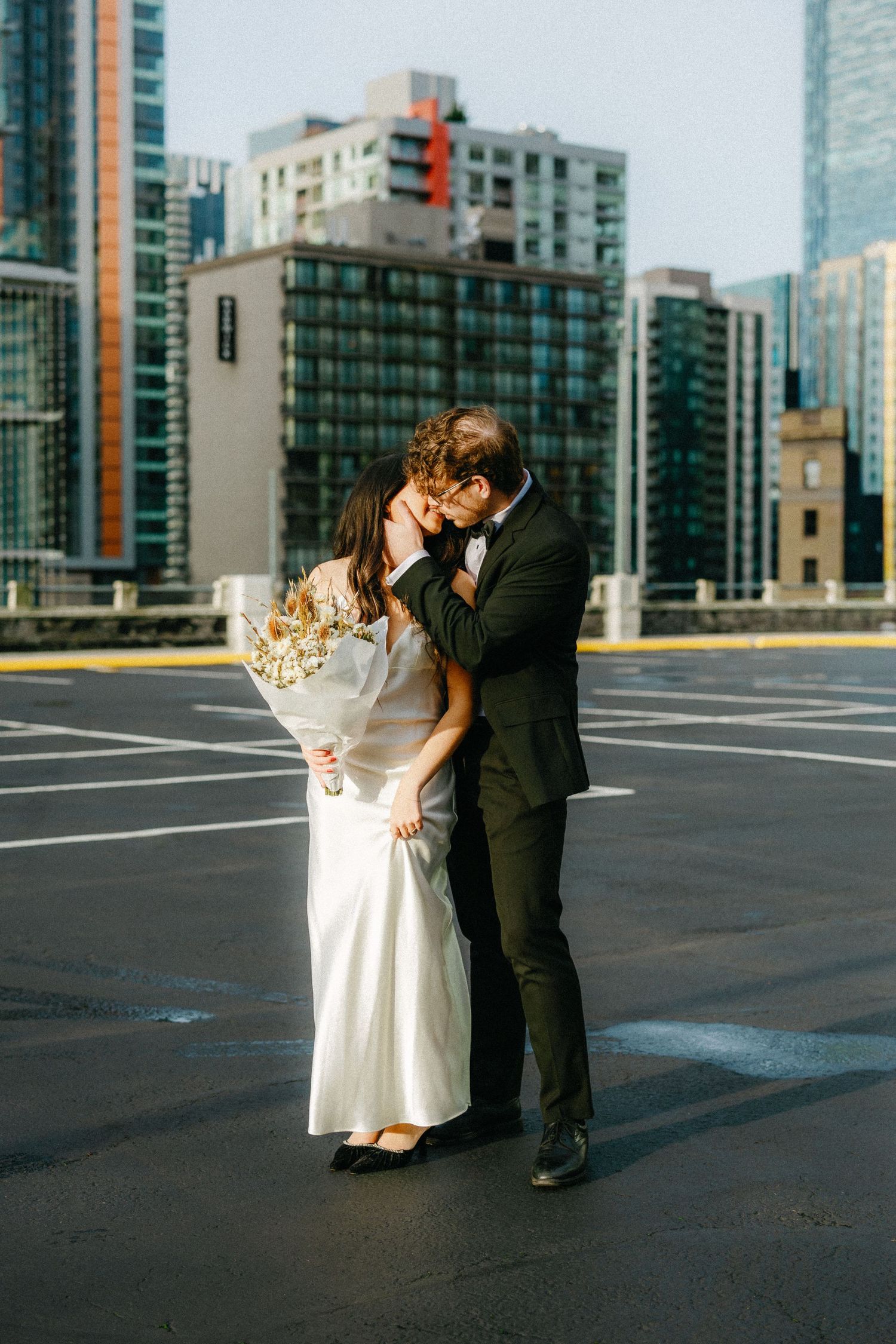 A couple shares a romantic moment on a rooftop parking lot with city buildings in the background.