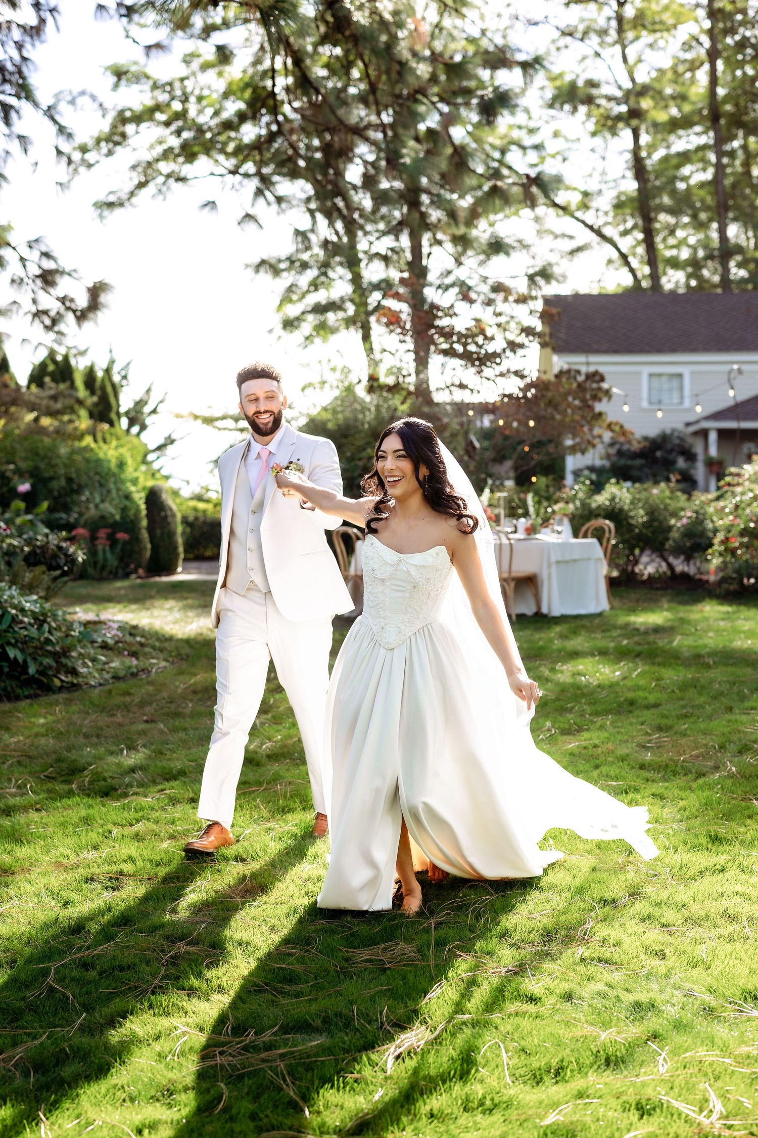 A couple dances playfully on a sunlit grassy lawn with trees and houses in the background.