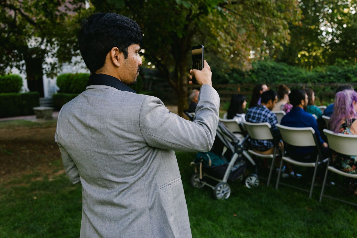 Someone in a gray jacket and black cap holds up a phone to take a photo at an outdoor gathering with tables and chairs.