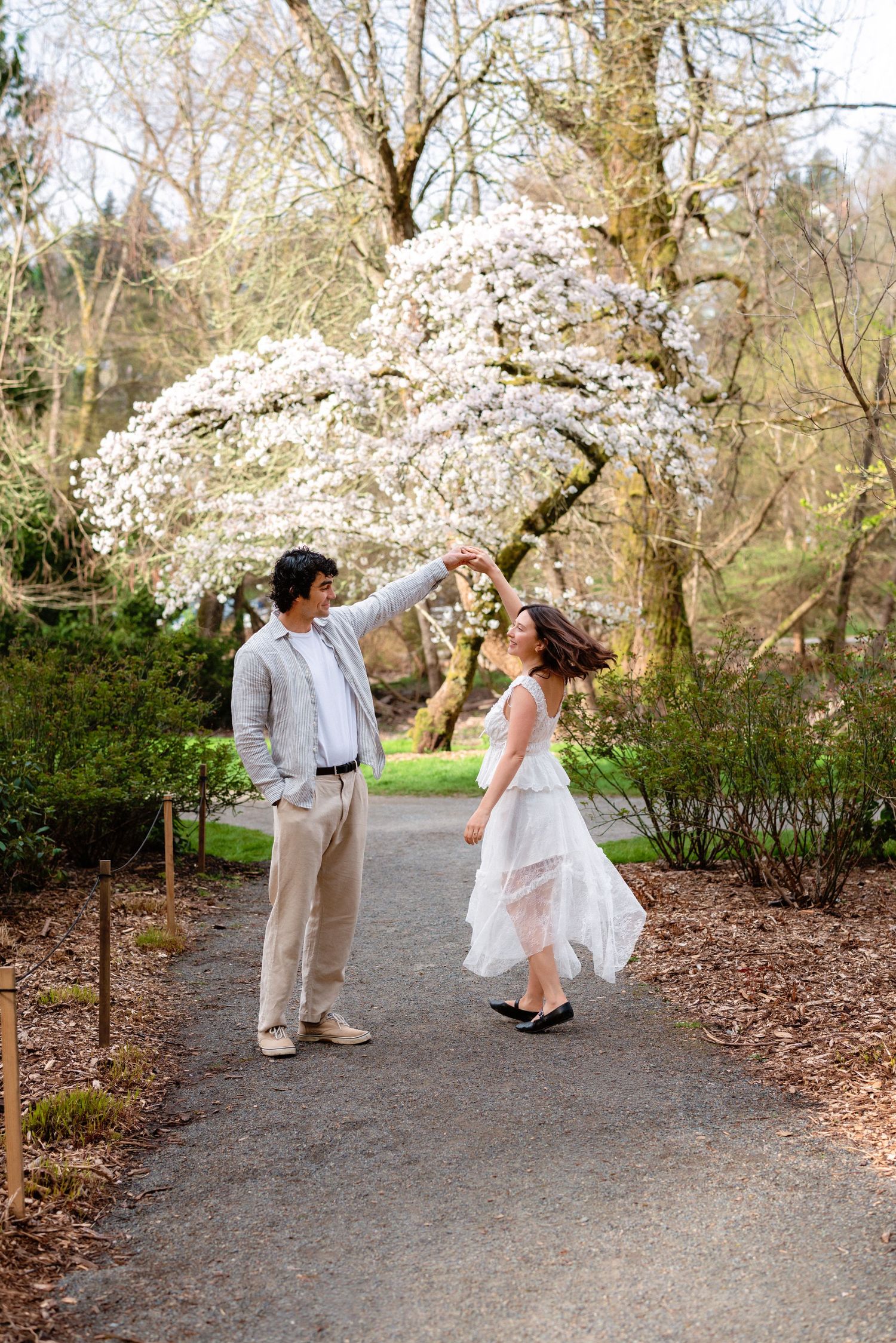 A couple dances playfully on a garden path beneath flowering cherry blossom trees.