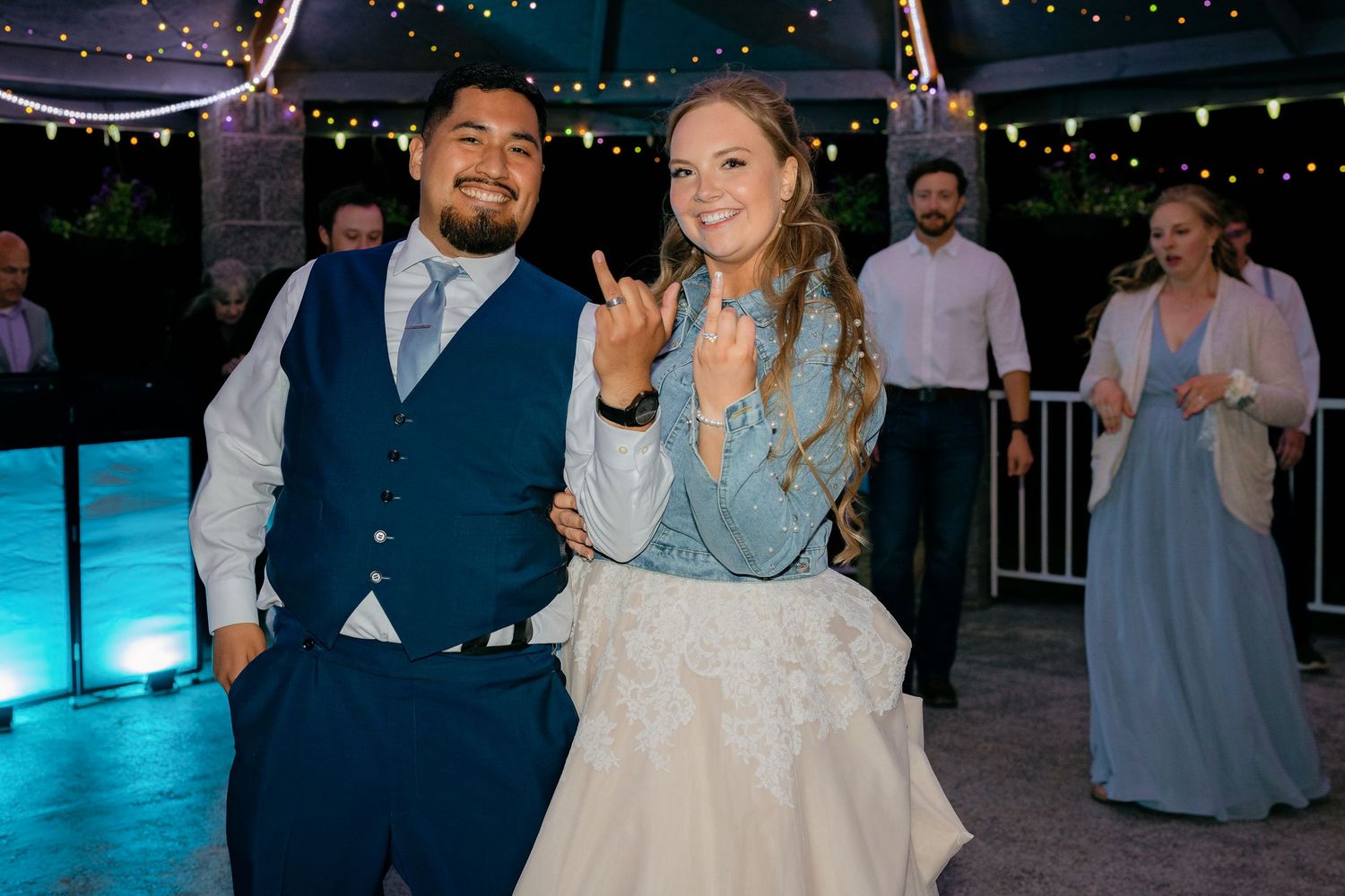 Smiling couple in wedding attire making rock on hand gestures on an outdoor dance floor at night.