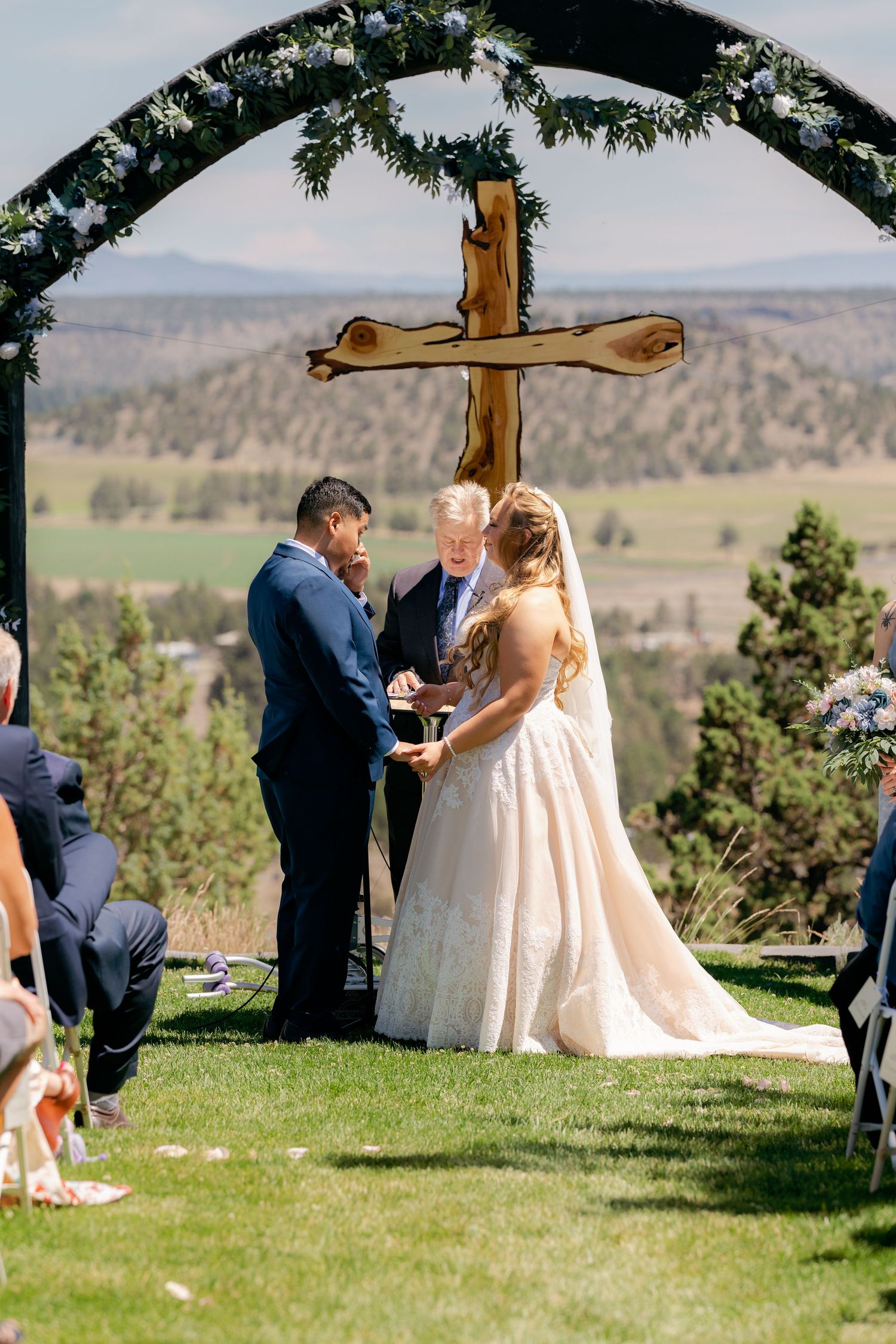 Bride and groom exchange vows under a wooden cross arch with scenic mountain views at outdoor wedding.