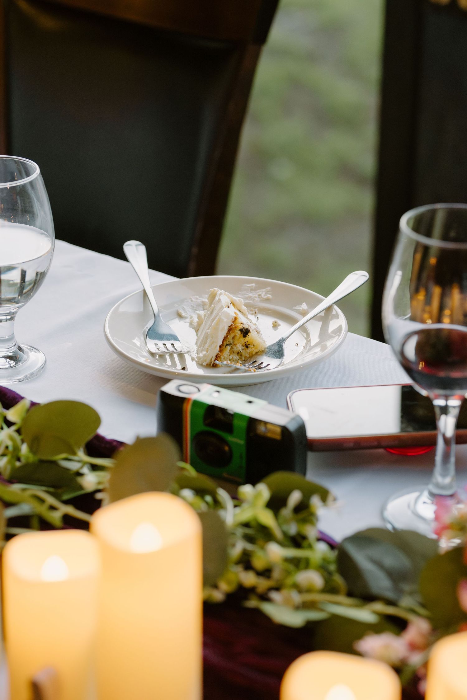Partially eaten slice of wedding cake on plate with forks, wine glasses, and candles on table.