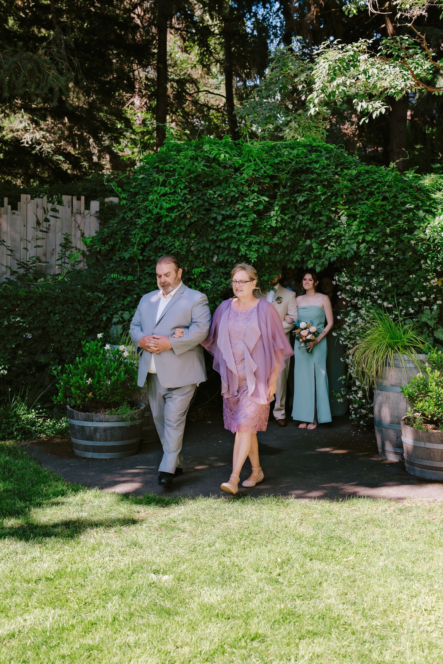 Mother escorted down aisle by son at outdoor garden wedding.