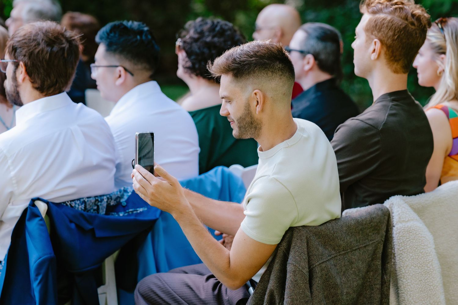 Wedding guest smiles while recording ceremony on smartphone at outdoor summer wedding.