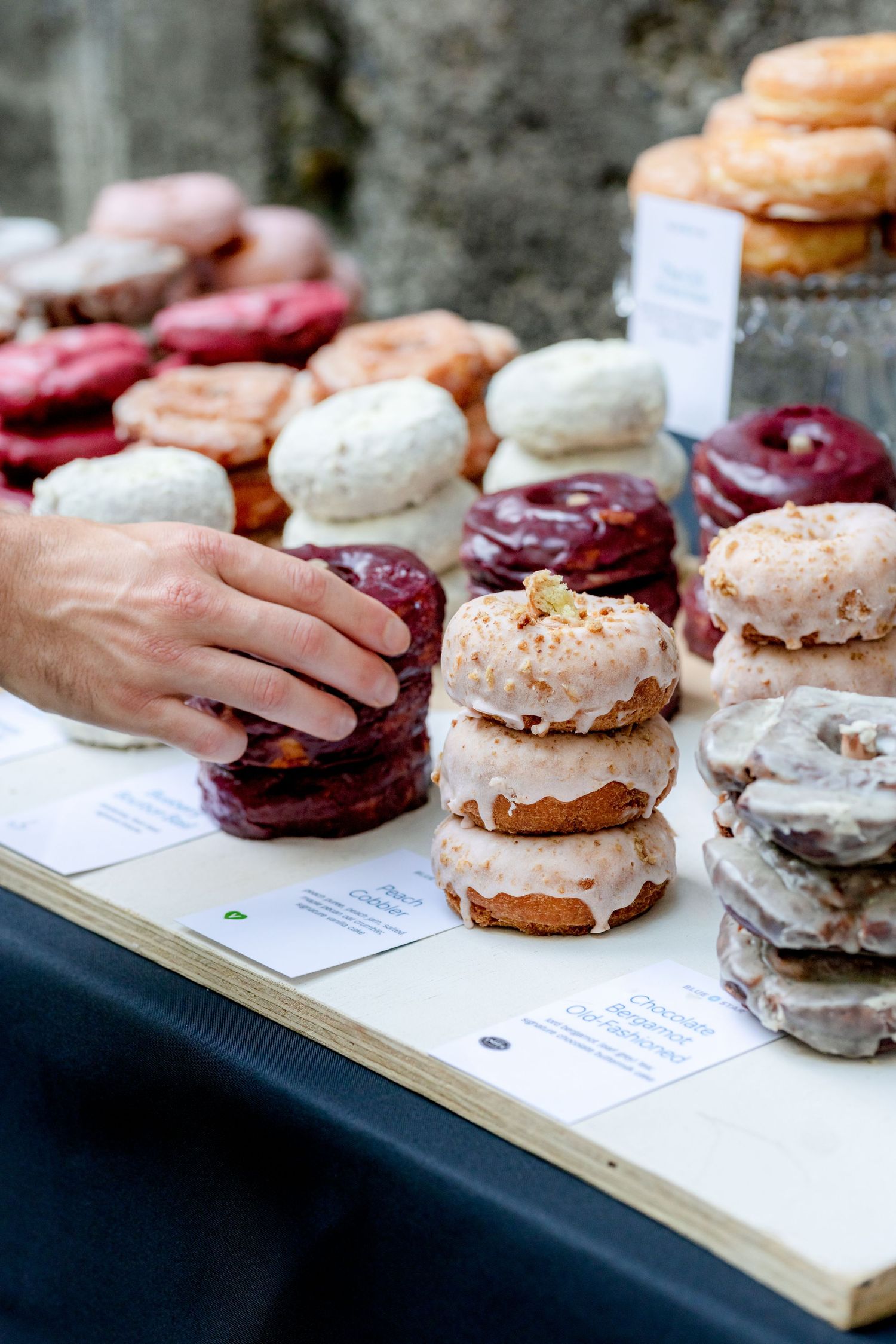 Assorted glazed donuts stacked on a wooden table at a market, with a hand reaching for a dark berry-frosted donut.