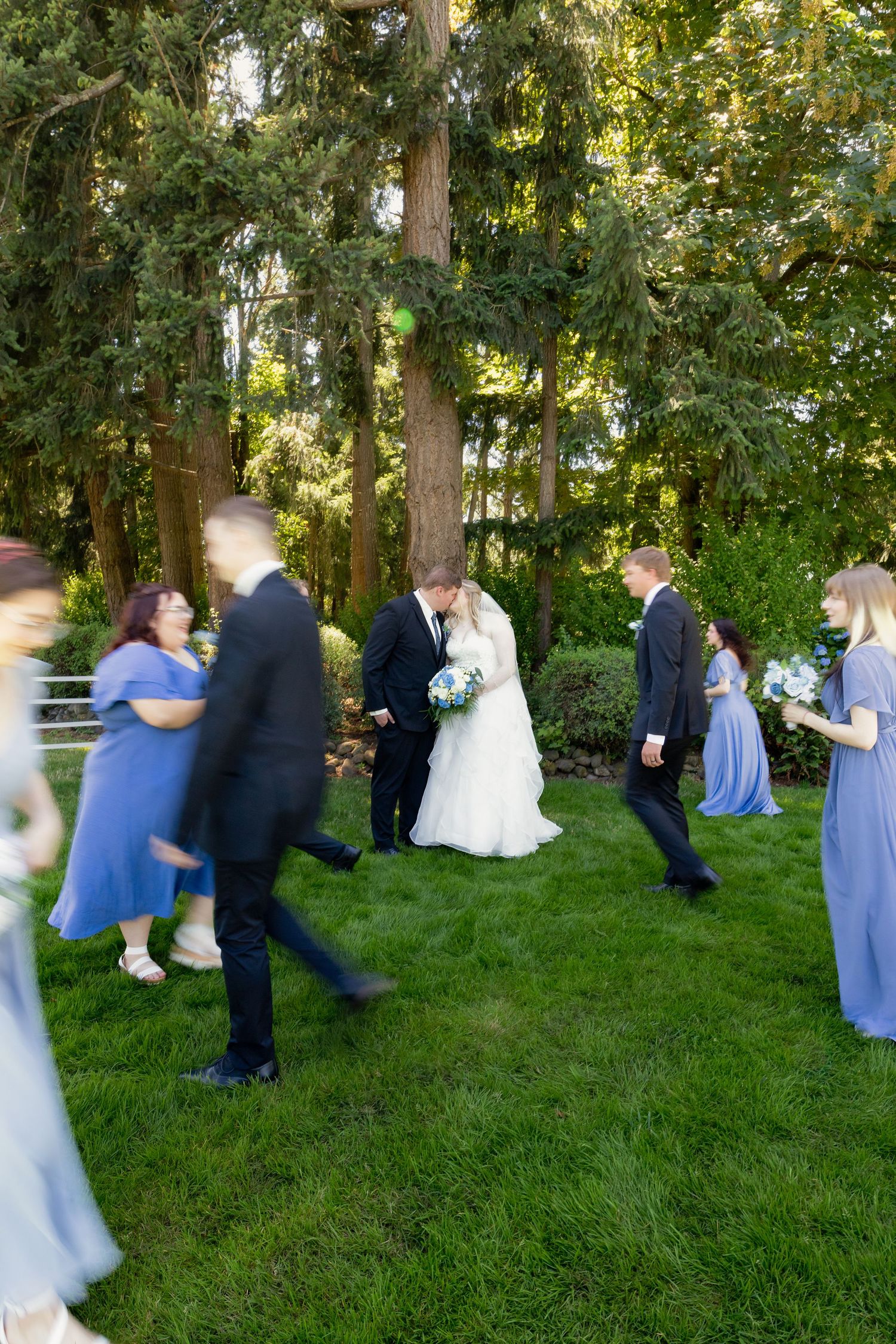 Wedding party in blue and black attire surrounds kissing bride and groom on a green lawn.