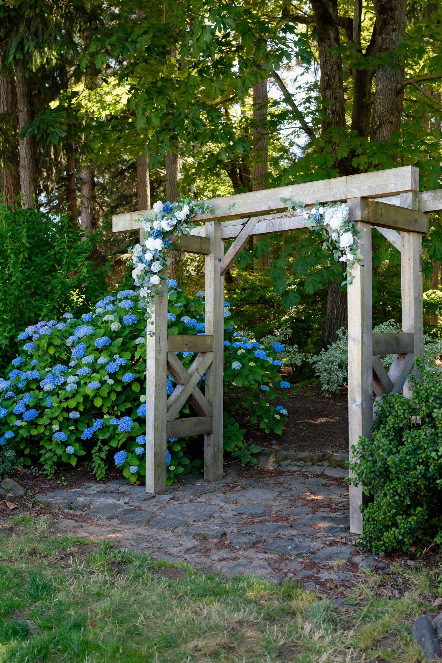 Wooden garden arbor decorated with white and blue flowers surrounded by hydrangeas and lush green trees.
