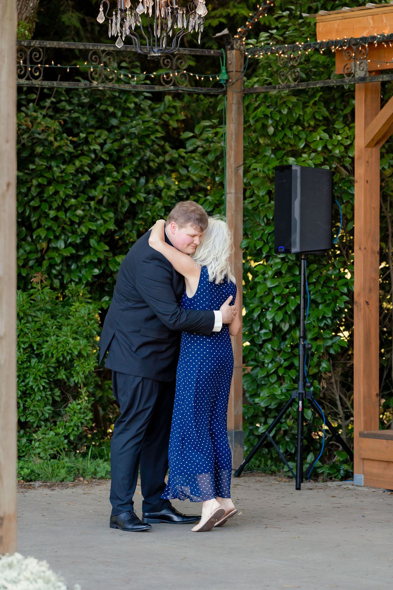 Groom in black suit shares tender dance with woman in blue polka dot dress at outdoor garden wedding reception.