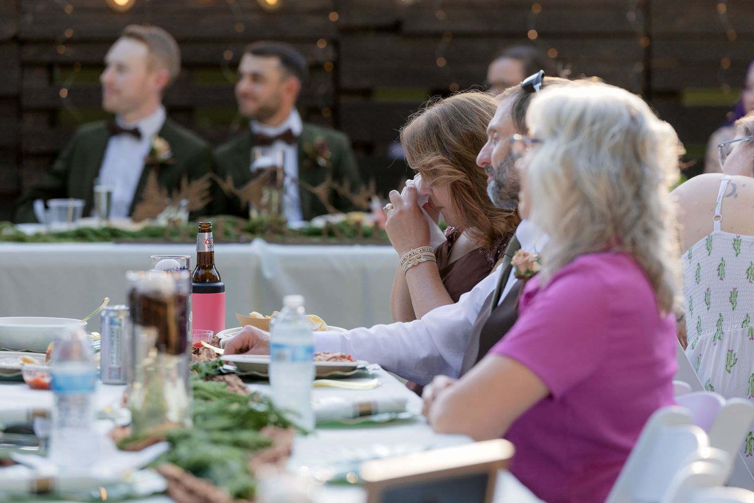 Wedding guests seated at a long decorated table listening emotionally to speeches during an outdoor reception.