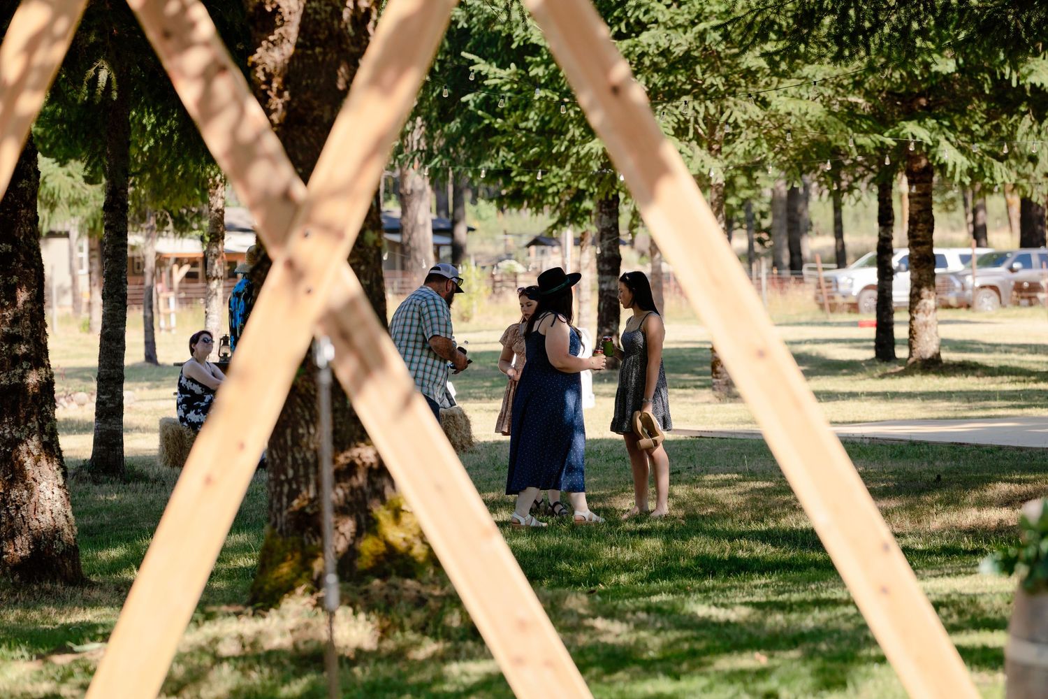 People gathered in a sunny park framed by wooden X-shaped structures among green trees.