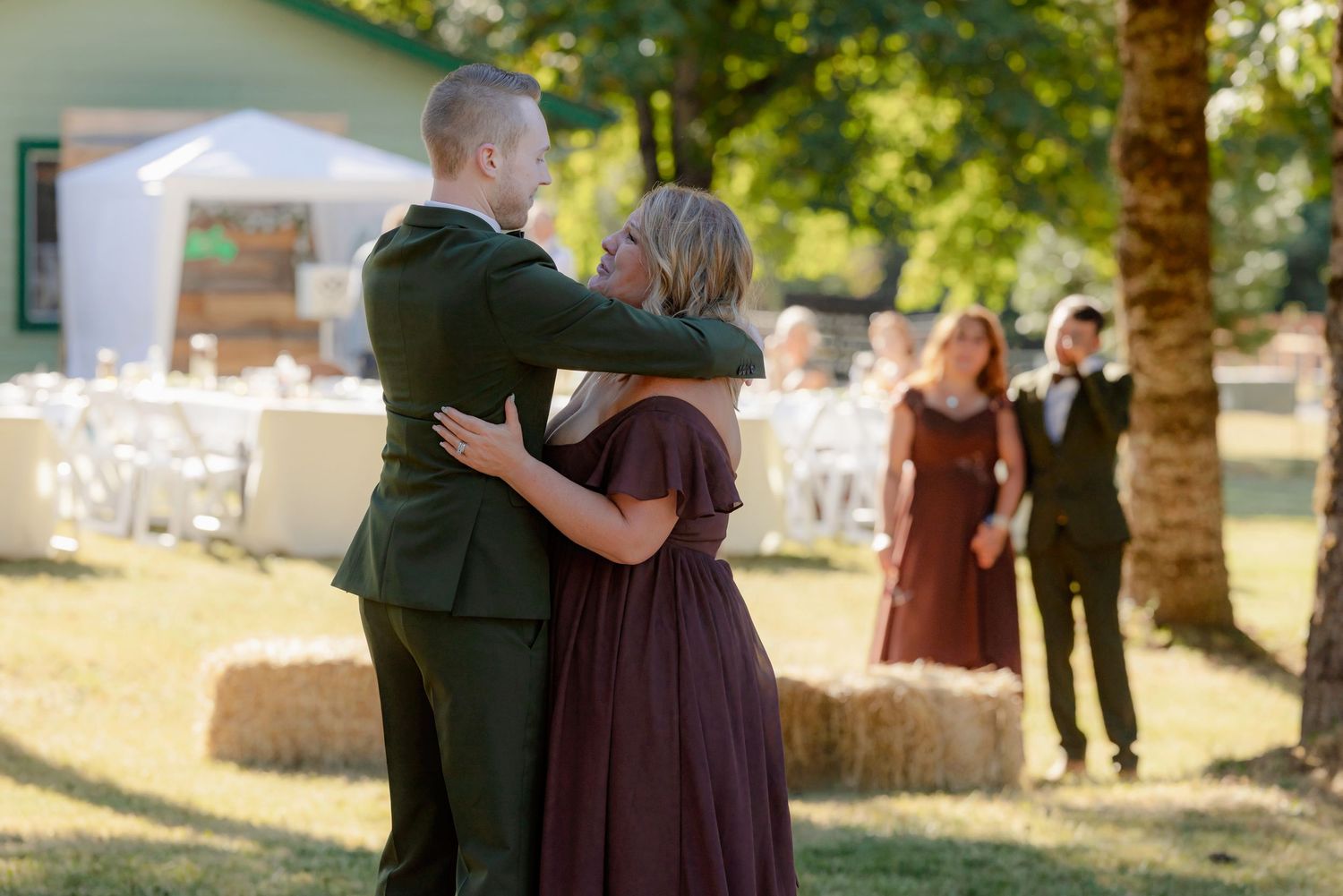 Groom embraces woman in burgundy dress during emotional parent dance at outdoor wedding reception.