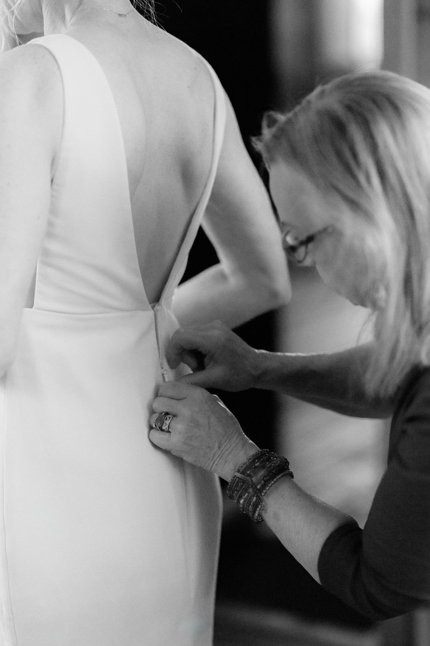 Black and white photo of a woman fastening the back of a bride's white wedding dress before the ceremony.