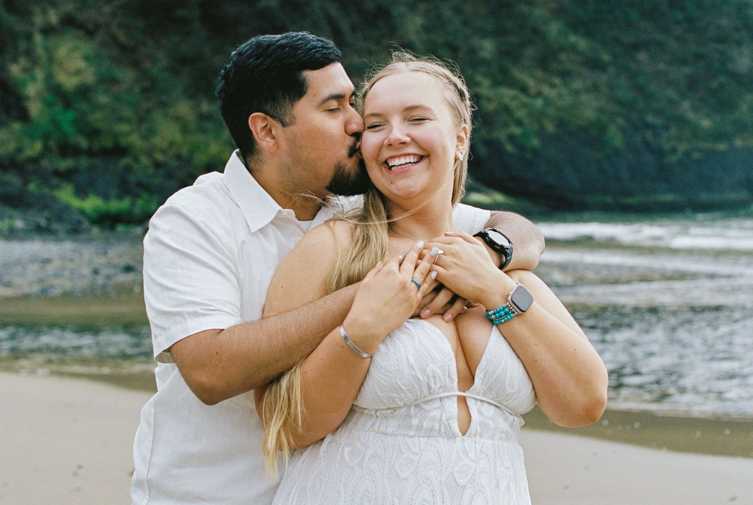 Man hugging woman from behind on a beach, both smiling, with lush green trees in the background.