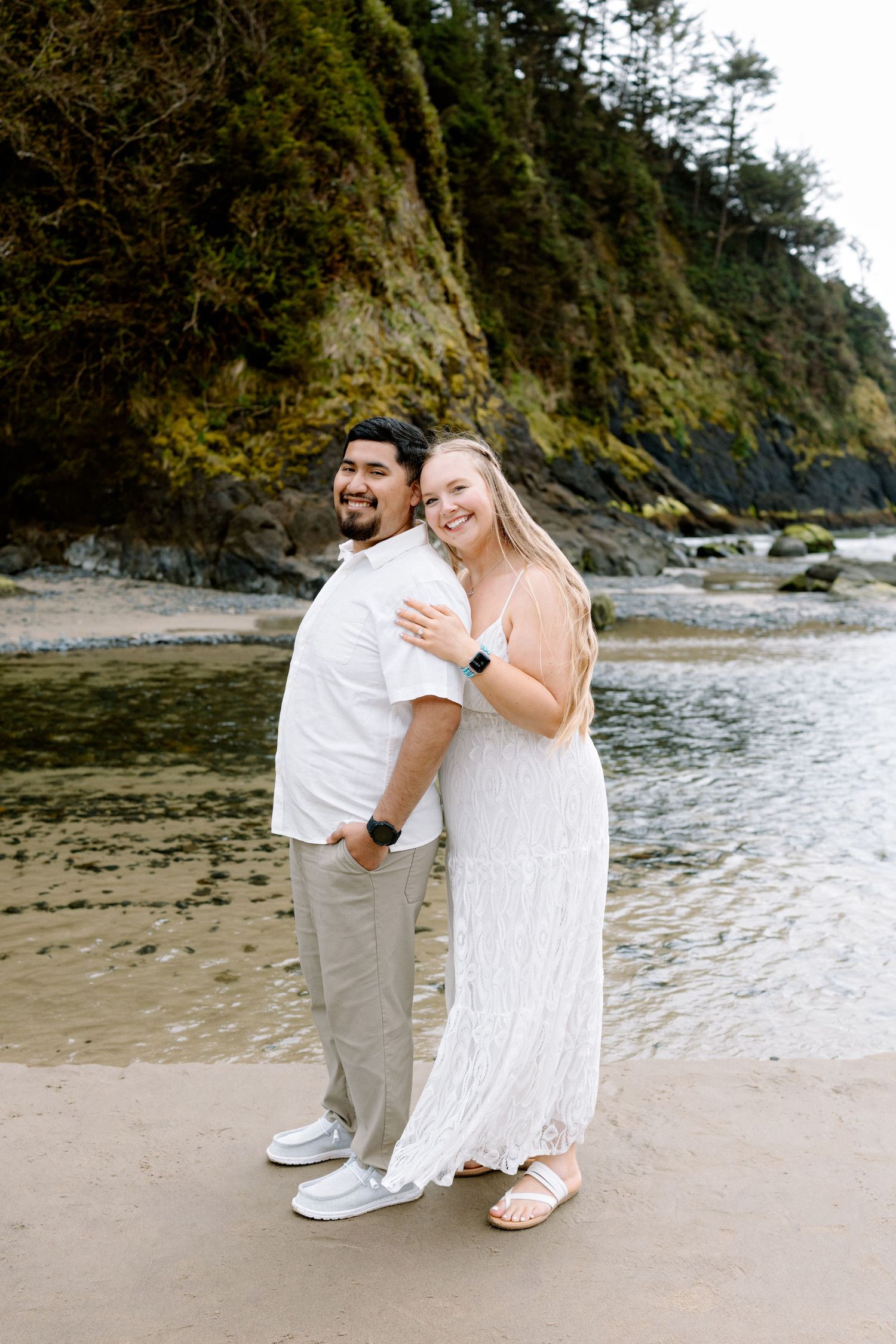 A smiling couple embraces on a sandy beach, wearing white outfits, with mossy cliffs and calm water in the background.