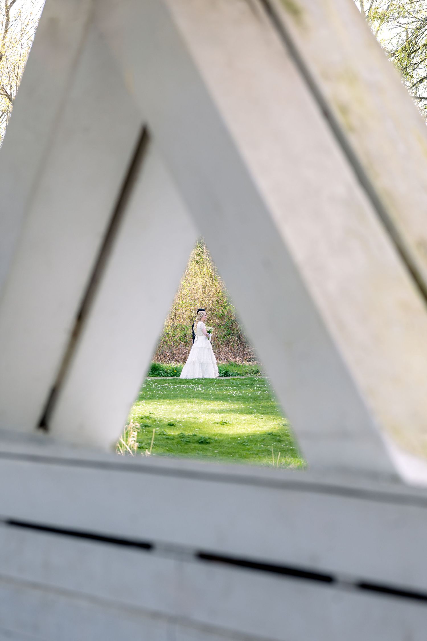 A bride in a white wedding dress seen through a triangular white wooden arch in a lush green garden.