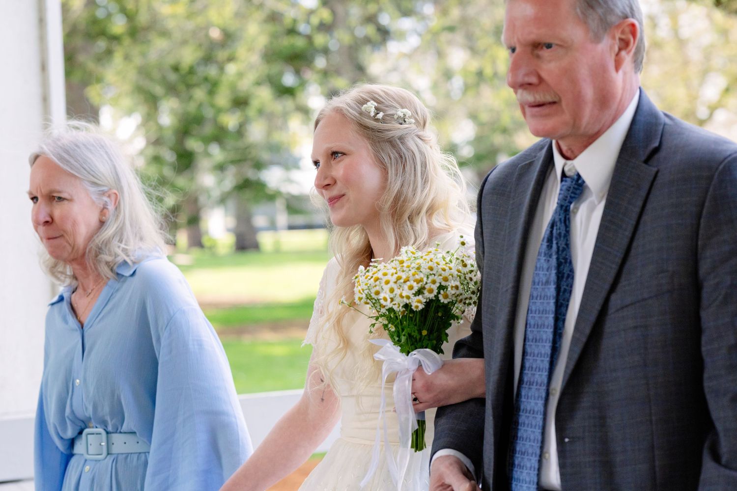 Young girl in white dress and tiara holds bouquet, escorted by elderly couple at outdoor wedding ceremony.