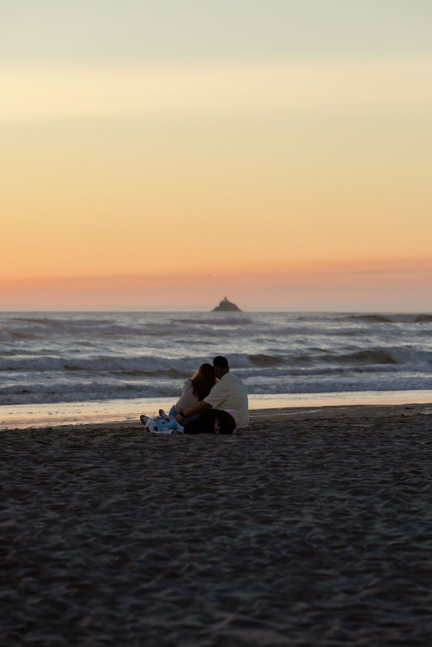 Couple sitting together on sandy beach watching romantic sunset over ocean waves with small island silhouette in distance.