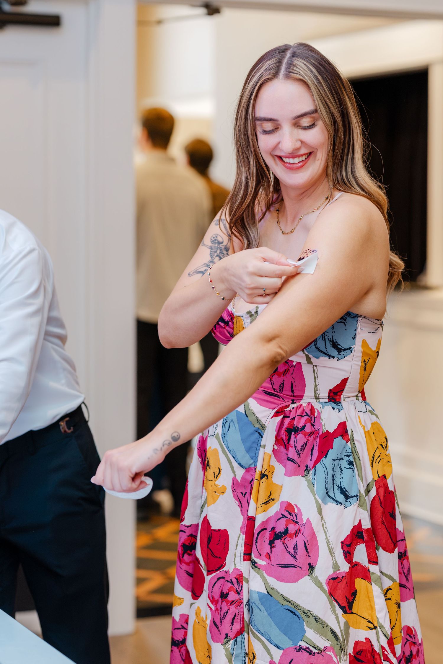 Smiling woman in a colorful floral dress applies perfume to her wrist at an indoor event.
