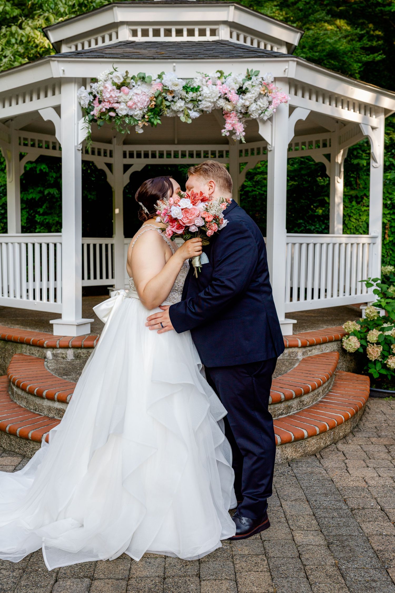 Bride and groom share a kiss holding a pink bouquet in front of a white floral gazebo on their wedding day.