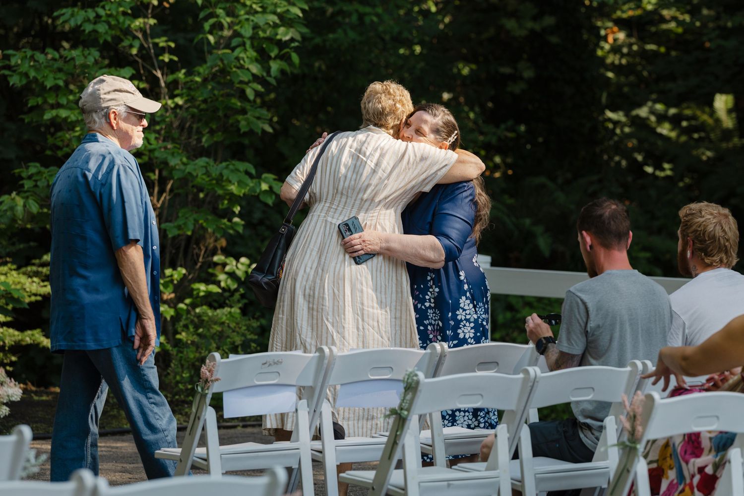 Two women share a warm hug at an outdoor wedding ceremony surrounded by white chairs and lush greenery.