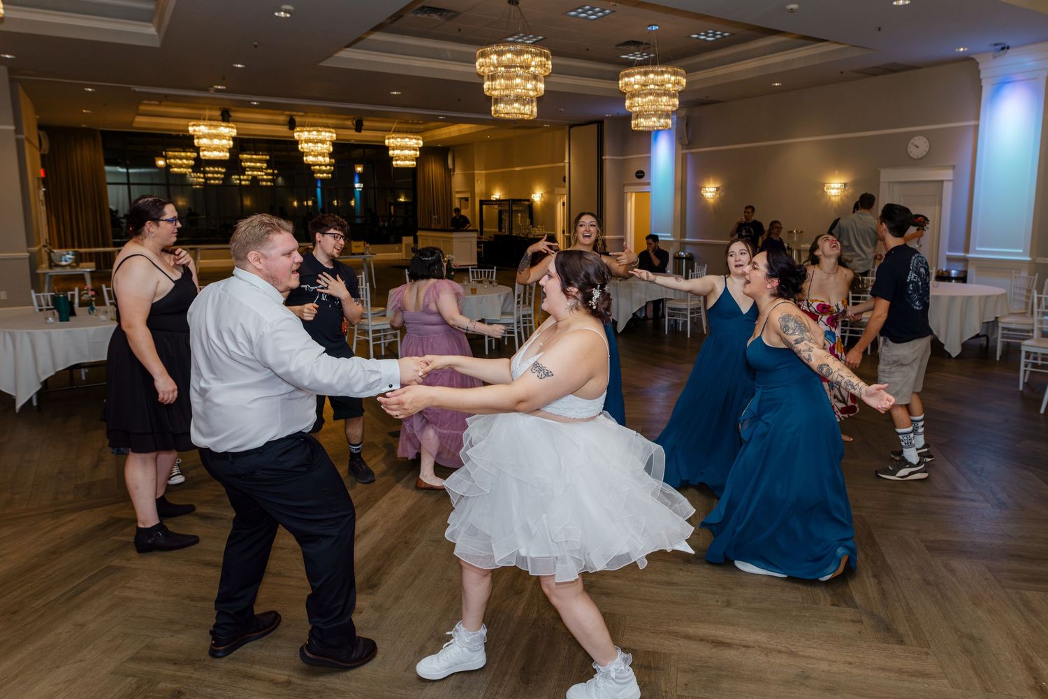 Bride in white dress dancing in a circle with guests including bridesmaids in blue at the reception.