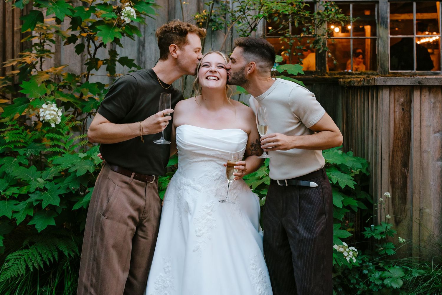 Bride in white strapless gown laughs as two men kiss her cheeks in a garden setting.