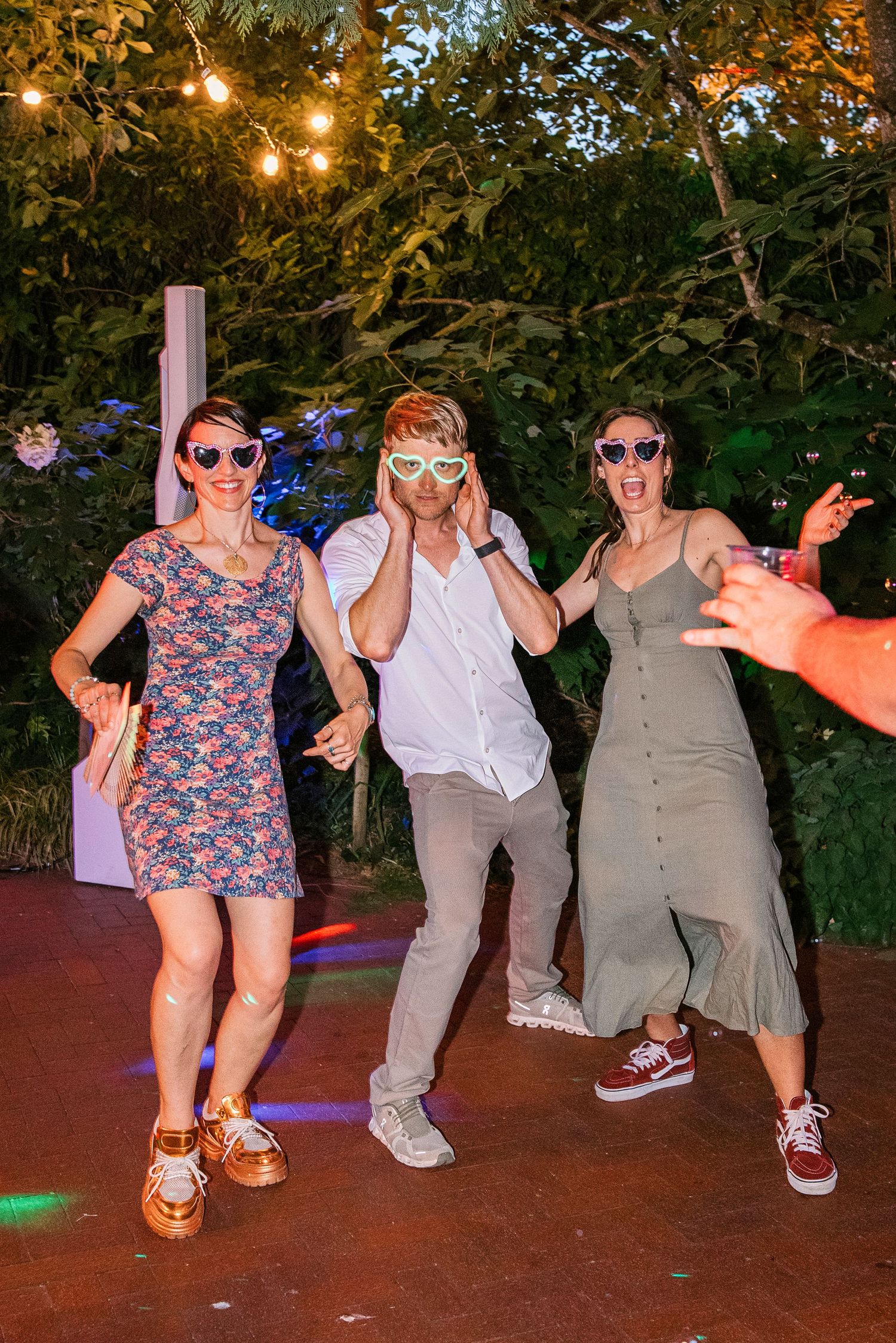 Three friends dancing outdoors at a party wearing fun sunglasses under string lights at night.